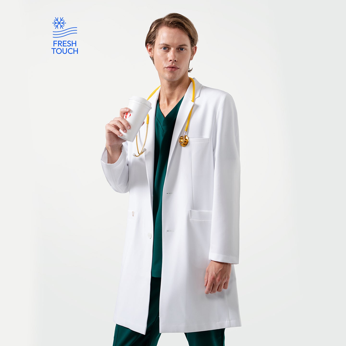 Image of Male doctor wearing a white lab coat over green scrubs with a yellow stethoscope around his neck, holding a white takeaway coffee cup